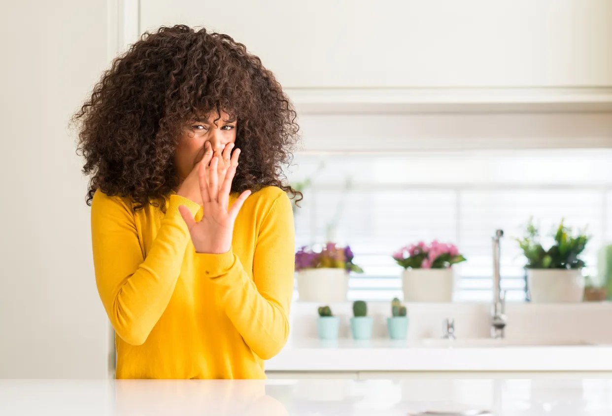 African American woman wearing a yellow sweater in the kitchen smelling something bad and holding her nose